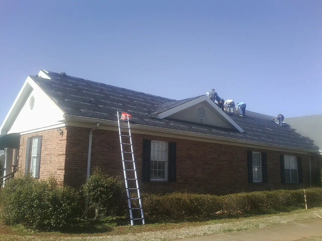 Crew tearing shingles off a brick ranch roof with a ladder set — Emergency Roof Repair work by our crew in Lone Tree